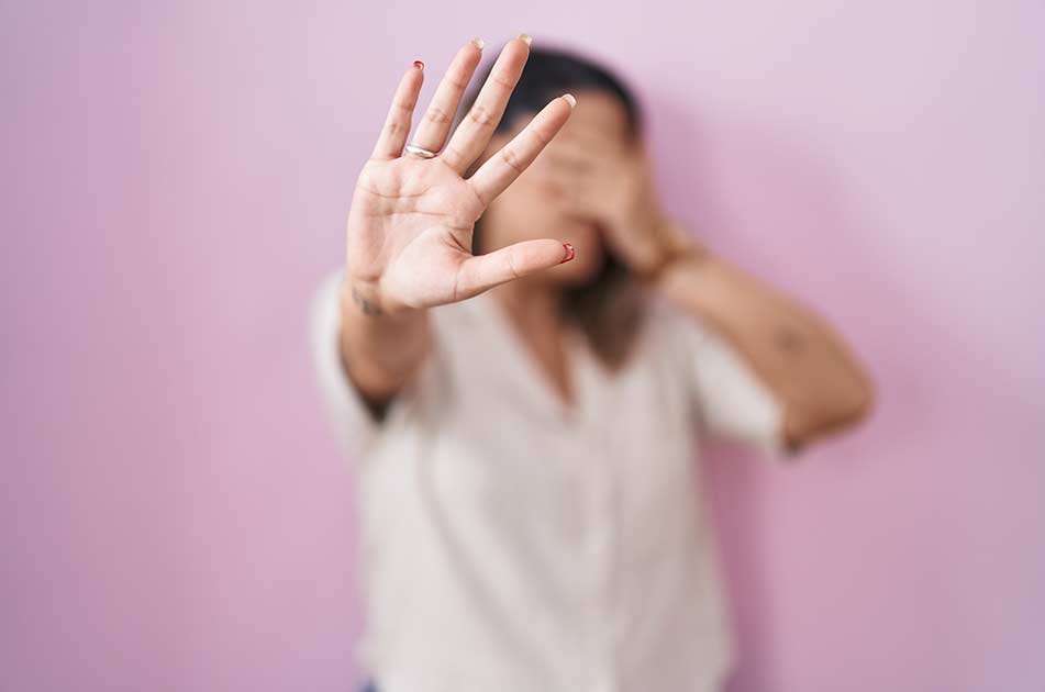 Eine Frau hält ihre Hand vor die Kamera, um ihr Gesicht zu verdecken. Sie scheint sich zu schützen oder abzulehnen. Der Hintergrund ist ein einfacher rosa Farbton, und die Person ist unscharf, was die Aufmerksamkeit auf die ausgestreckte Hand lenkt.